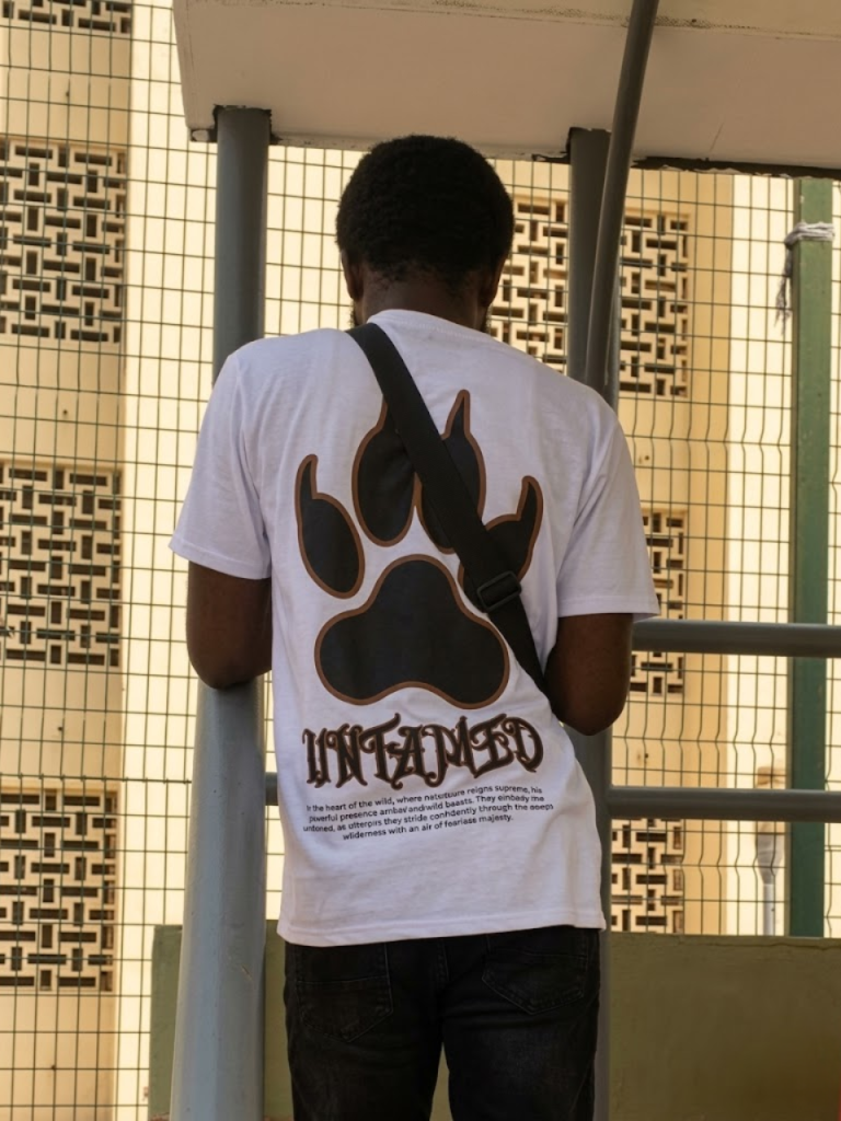 A young male wearing the white colorway of the Untamed Tee from DripAfrika, showing the bold paw graphic at the back of the shirt