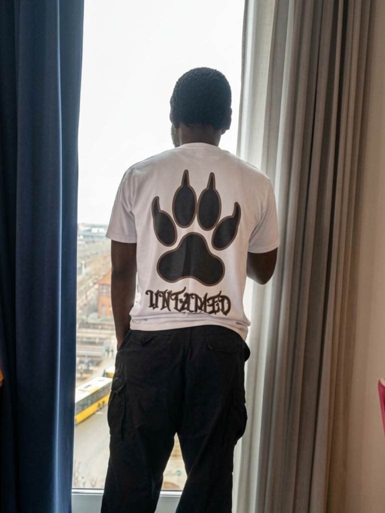 A young male wearing the white colorway of the Untamed Tee from DripAfrika, showing the bold paw graphic at the back of the shirt